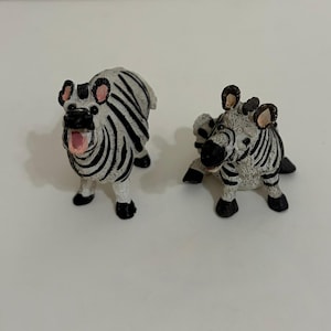 May include: Two small zebra figurines with black and white striped patterns. One zebra stands with its mouth open, revealing a pink interior, while the other zebra sits in a resting position. Both figures feature black hooves and ears.