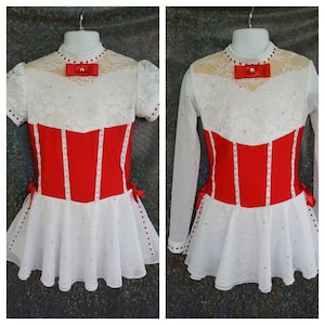 May include: A white figure skating dress with a red bodice and a white lace overlay. The bodice has a red bow and is decorated with rhinestones. The skirt is layered and has a white lace trim.