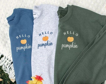 Könnte beinhalten: Drei Sweatshirts mit dem Text "Hello Pumpkin" und einer kleinen Kürbisgrafik. Die Sweatshirts sind in Blau, Grau und Grün. Die Sweatshirts liegen auf einer weißen Decke mit Herbstblättern und einem großen orangefarbenen Kürbis.