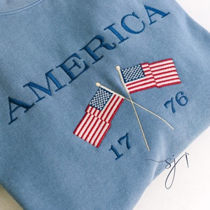 May include: A blue sweatshirt with the word "AMERICA" embroidered in white. Two American flags are embroidered below the word, crossed at their poles. The numbers "17" and "76" are embroidered below the flags.