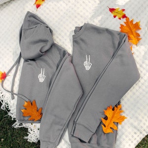 May include: Two gray sweatshirts with a white skeleton hand peace sign design. The sweatshirts are laid out on a white blanket with fall leaves.