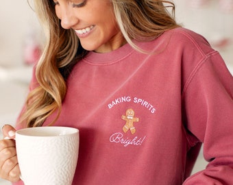 Może przedstawiać: Kobieta w różowej bluzie z napisem "Baking Spirits Bright!" i grafiką piernikowego ludzika. Trzyma biały kubek.