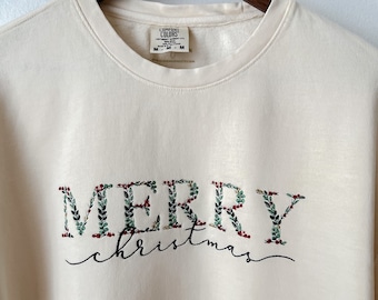 Może przedstawiać: Kremowa bluza z haftem "Merry Christmas" w kolorze zielonym, czerwonym i białym. Litery są obramowane zielonymi liśćmi i czerwonymi jagodami.
