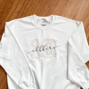 May include: White sweatshirt with a gold embroidered number 18 and the word "eighteen" in cursive script.