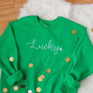 May include: Green sweatshirt with the word "Lucky" in white cursive lettering and a small shamrock. The sweatshirt is surrounded by gold coins on a wooden surface.