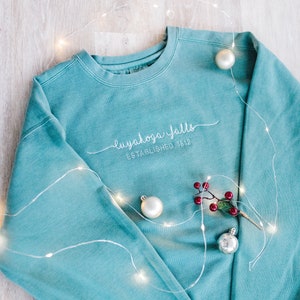 May include: A teal blue sweatshirt with white embroidered text that reads "Cuyahoga Falls Established 1812". The sweatshirt is surrounded by white fairy lights and red berries.