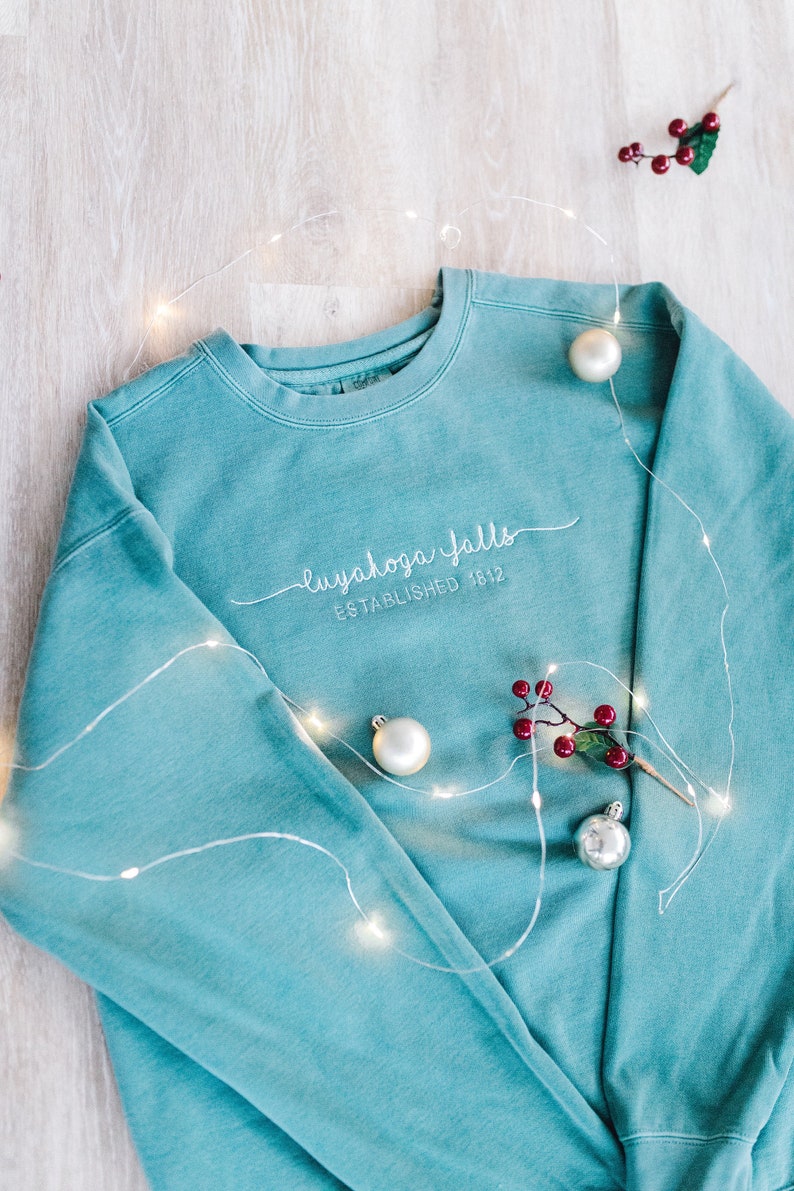 May include: A teal blue sweatshirt with white embroidered text that reads "Cuyahoga Falls Established 1812". The sweatshirt is surrounded by white fairy lights and red berries.