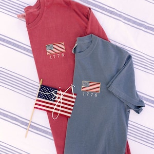 May include: Two t-shirts with a small American flag and the year 1776 embroidered on the front. One shirt is red and the other is blue.