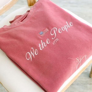 May include: A red sweatshirt with the text "We the People 1776" embroidered in white. A small American flag is embroidered above the text.