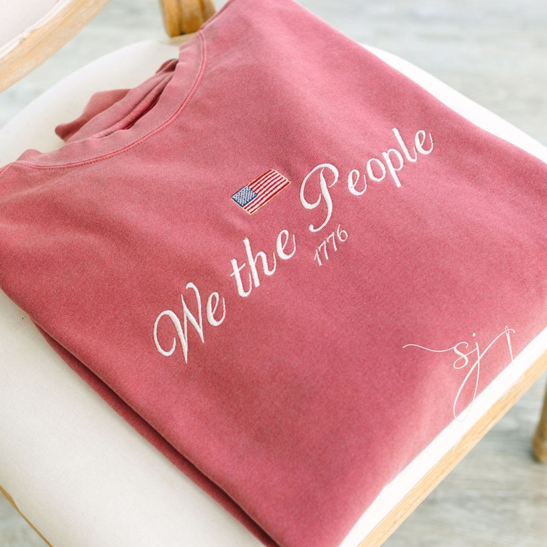 May include: A red sweatshirt with the text "We the People 1776" embroidered in white. A small American flag is embroidered above the text.