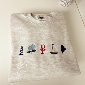 May include: A light gray heathered crewneck sweatshirt featuring five small embroidered icons across the chest: a blue and white lighthouse, a blue hydrangea flower, a red lobster, a sailboat, and a dark blue silhouette of the state of Maine.
