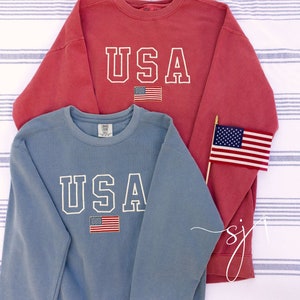 May include: Two sweatshirts, one red and one blue, each with "USA" in white block letters and a small American flag. The sweatshirts are displayed on a striped background, with a small American flag on a stick.