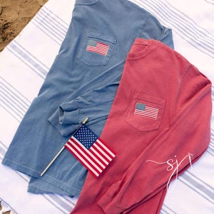 May include: Two long-sleeved shirts, one blue and one red, each with a pocket featuring an embroidered American flag. A small American flag on a stick rests between the shirts. The shirts are on a striped surface.