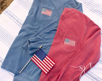 May include: Two long-sleeved shirts, one blue and one red, each with a pocket featuring an embroidered American flag. A small American flag on a stick rests between the shirts. The shirts are on a striped surface.