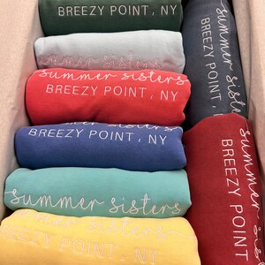 May include: A stack of folded t-shirts in various colours, each with the text "summer sisters" and "BREEZY POINT, NY" embroidered on them.