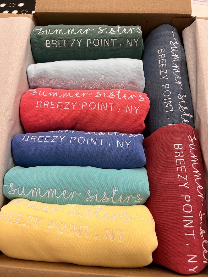 May include: A stack of folded t-shirts in various colours, each with the text "summer sisters" and "BREEZY POINT, NY" embroidered on them.
