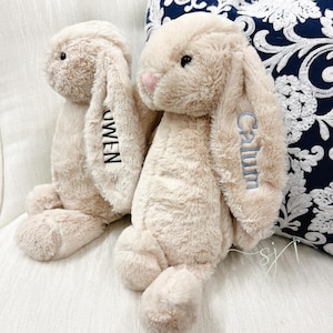 May include: Two plush beige bunny rabbits with long floppy ears. One ear has the name "Owen" embroidered in black, and the other has "Calum" embroidered in blue. The bunnies have pink noses and are sitting on a white surface.