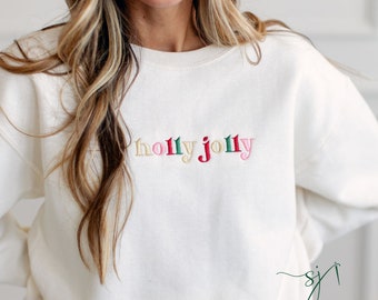 May include: Cream-colored sweatshirt with the embroidered text "holly jolly" in various colors. The word "holly" is in gold, red, and green, while "jolly" is in green and pink. The sweatshirt has a crew neck and long sleeves.