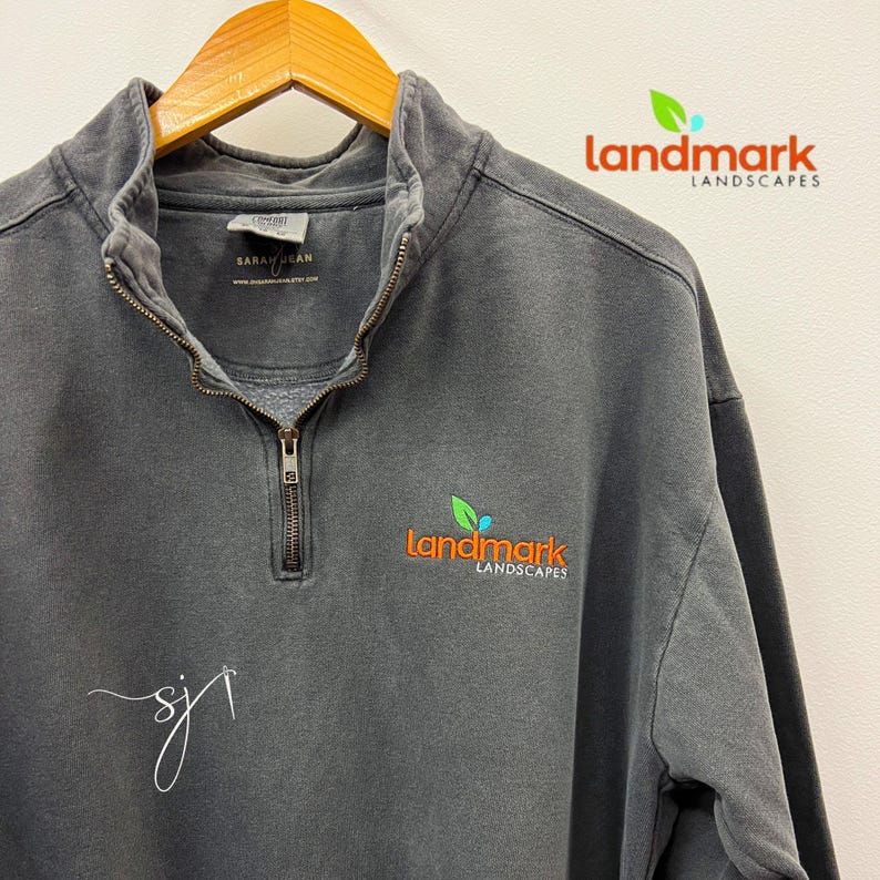 May include: A dark gray, 1/4 zip sweatshirt with the text "landmark landscapes" embroidered on the right chest in orange, green, and blue. The sweatshirt has a zipper and a small tag on the inside neck that says "Sarah Jean".