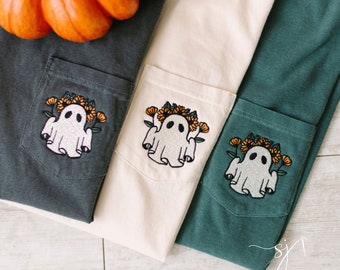 May include: Three t-shirts with a ghost embroidered on the pocket. The ghost is wearing a crown of flowers. The t-shirts are in different colors: dark gray, cream, and teal.