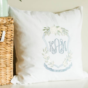 May include: White decorative pillow with an embroidered monogram in light blue. The monogram is surrounded by a floral wreath and a banner with a date. The pillow is placed next to a woven basket.