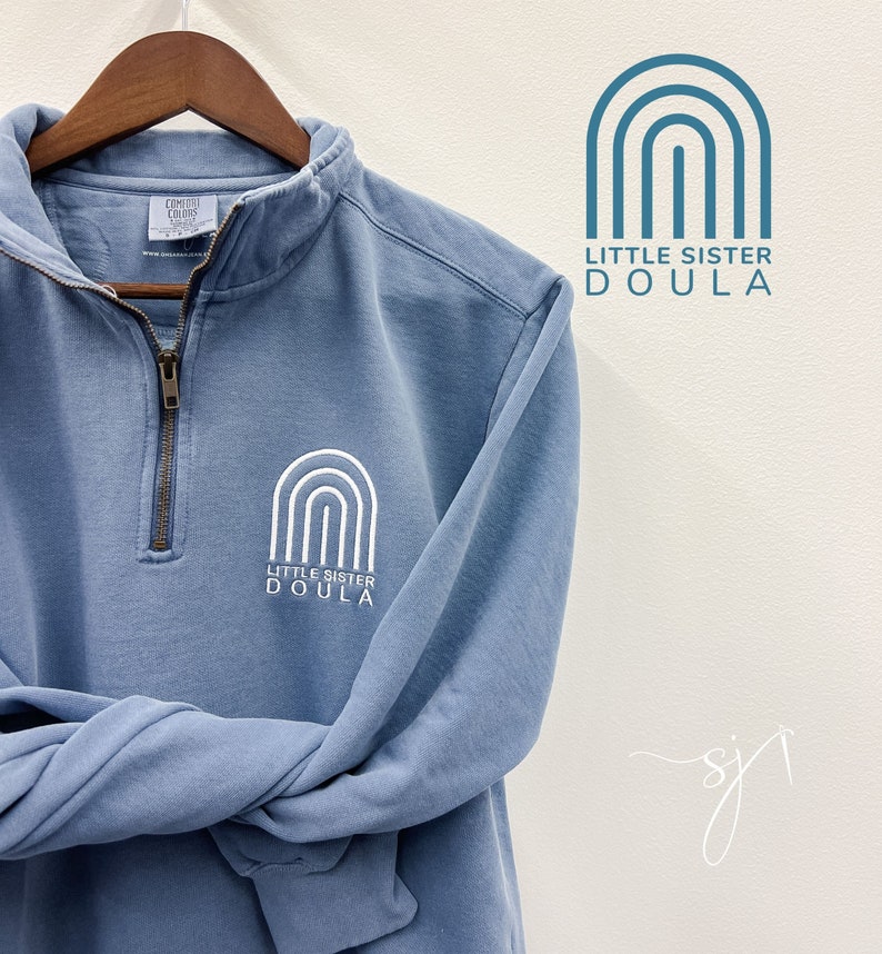 May include: A light blue sweatshirt with a white rainbow graphic and the text "Little Sister Doula".