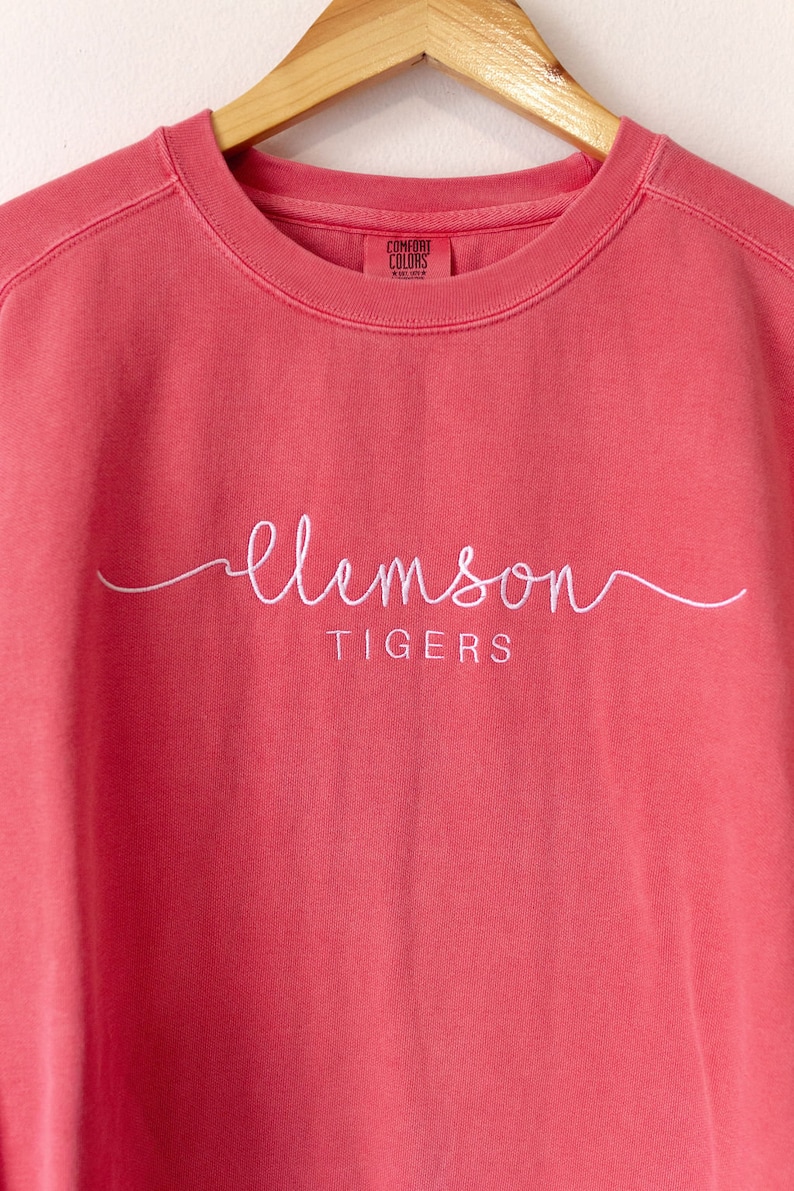 May include: A coral pink sweatshirt with the words "Clemson Tigers" embroidered in white.