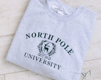 Może przedstawiać: Szara bluza z zielonym haftem, na którym widnieje napis "North Pole University" oraz herb z jeleniem i tekstem "Est. 1862".