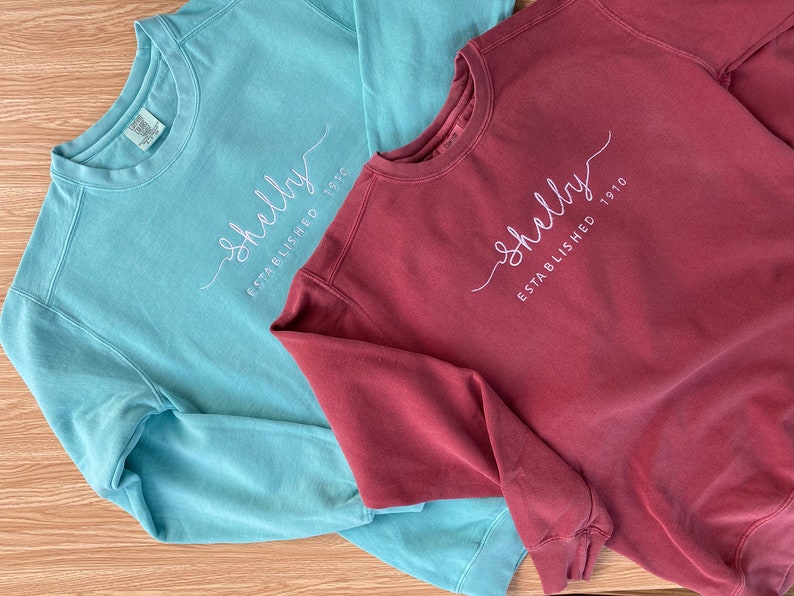 May include: Two sweatshirts, one teal blue and one maroon, with the text "Shelly Established 1910" embroidered on the front.