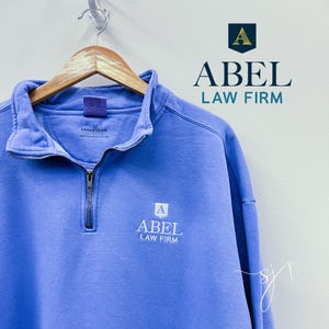 May include: A light blue quarter-zip sweatshirt with the logo "ABEL LAW FIRM" embroidered on the front. The logo is white with a small gold "A" in a shield shape.