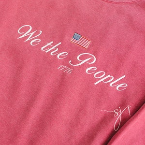 May include: A pink long-sleeved t-shirt with the text "We the People 1776" in white cursive lettering and a small American flag embroidered on the left side of the chest.