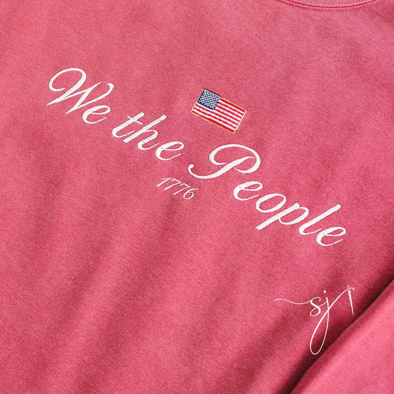May include: A pink long-sleeved t-shirt with the text "We the People 1776" in white cursive lettering and a small American flag embroidered on the left side of the chest.