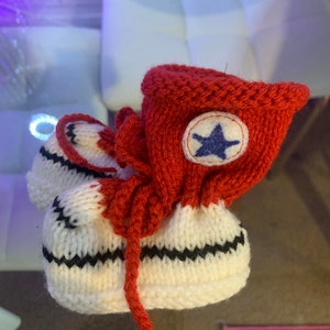 Puede incluir: Botines de bebé tejidos a mano al estilo de zapatillas altas. Los botines son principalmente blancos con rayas en zigzag negras y detalles rojos. Un emblema de estrella azul está en el puño rojo del tobillo. Los botines están diseñados para parecerse a zapatillas en miniatura.