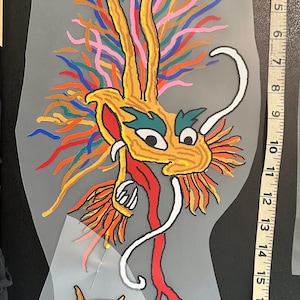 Könnte beinhalten: Bunte Drachenillustration mit gelbem Kopf, roter Zunge und mehrfarbigen Akzenten. Der Drache hat unten eine Sternform. Das Bild befindet sich auf einem transparenten, unregelmäßig geformten Hintergrund. Ein Maßband befindet sich auf der rechten Seite.