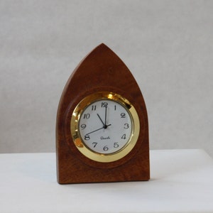 May include: A wooden clock with a gold-colored frame and a white face. The clock is shaped like a pointed arch and has a simple design.