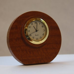May include: A round wooden clock with a gold rim and white face. The clock is sitting on a white surface.