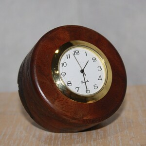 May include: A small, round, wooden clock with a white face and gold trim. The clock has black hands and black numbers. The clock is sitting on a wooden surface.