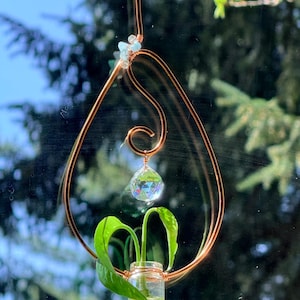 May include: Copper wire plant hanger with a glass vial holding a small plant. The hanger features a teardrop shape with a spiral and a faceted crystal. Small blue beads add detail to the top.