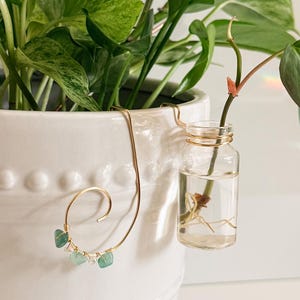 May include: A white ceramic planter with a trailing green plant. A gold-colored wire plant hanger with three small, light green gemstones is attached to the planter. A small glass vial with a plant cutting is suspended from the hanger.
