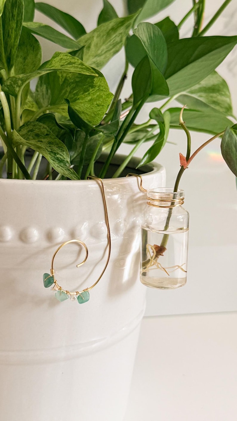 May include: A white ceramic planter with a trailing green plant. A gold-colored wire plant hanger with three small, light green gemstones is attached to the planter. A small glass vial with a plant cutting is suspended from the hanger.