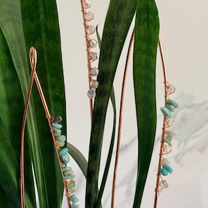 May include: Copper wire plant stakes adorned with small, colorful gemstones. The stakes are inserted into a green plant with long, pointed leaves. The gemstones are various shades of blue, clear, and white.
