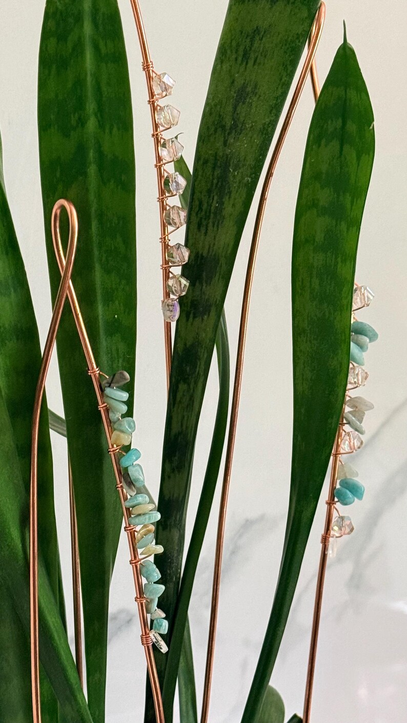 May include: Copper wire plant stakes adorned with colorful gemstones and clear crystals. The stakes are placed in a snake plant with long, green leaves. The gemstones are various sizes and shapes, adding a decorative touch to the plant.