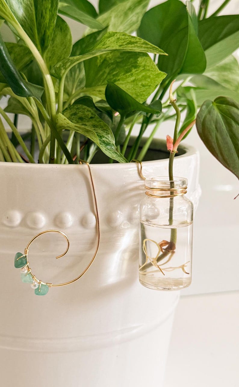 May include: A white ceramic planter with green leafy plants and a small glass propagation vase. A gold-colored wire plant hanger with green and clear beads is attached to the planter.
