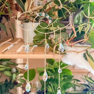 May include: A decorative gold-colored metal mobile with a cloud-like design, adorned with clear, iridescent crystal teardrops hanging from delicate chains. The mobile also features green and clear beads. The background includes lush green plants.