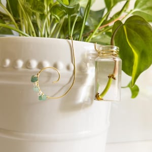 May include: A gold-colored plant propagation station with a clear glass vial and a decorative wire design. The wire features a spiral with small green and white stones. The station is attached to a white ceramic pot with green plants.