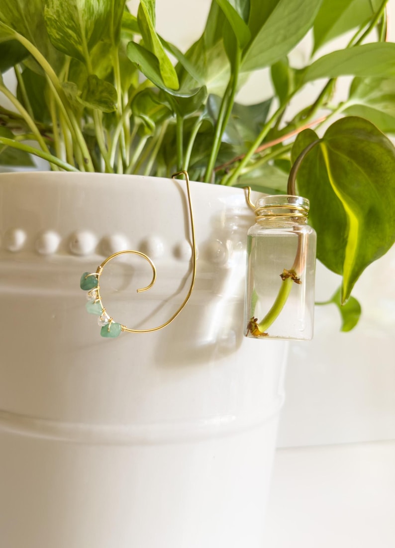 May include: A gold-colored plant propagation station with a clear glass vial and a decorative wire design. The wire features a spiral with small green and white stones. The station is attached to a white ceramic pot with green plants.