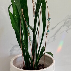 May include: A potted snake plant with copper wire accents adorned with small, light blue and clear gemstones. The plant is in a white ceramic pot with a beaded design. The background is a white marble wall.