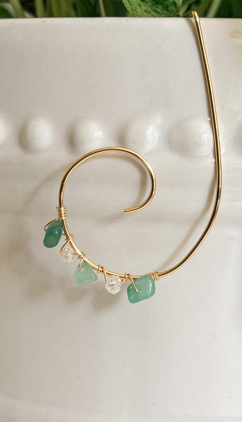 May include: A gold-colored metal spiral-shaped bookmark. The bookmark is adorned with small, polished green and clear gemstones. The gemstones are wire-wrapped onto the spiral, adding a touch of elegance to this unique accessory.