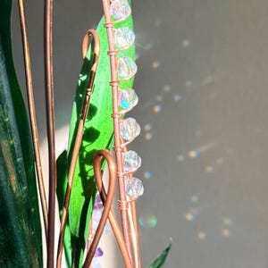 May include: A close-up of a copper wire plant stake with clear, iridescent beads attached to a green leaf. The stake is intertwined with other copper wires and green leaves. The background is a neutral gray.