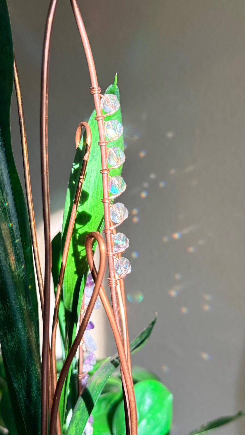 May include: A close-up of a copper wire plant stake with clear, iridescent beads attached to a green leaf. The stake is intertwined with other copper wires and green leaves. The background is a neutral gray.